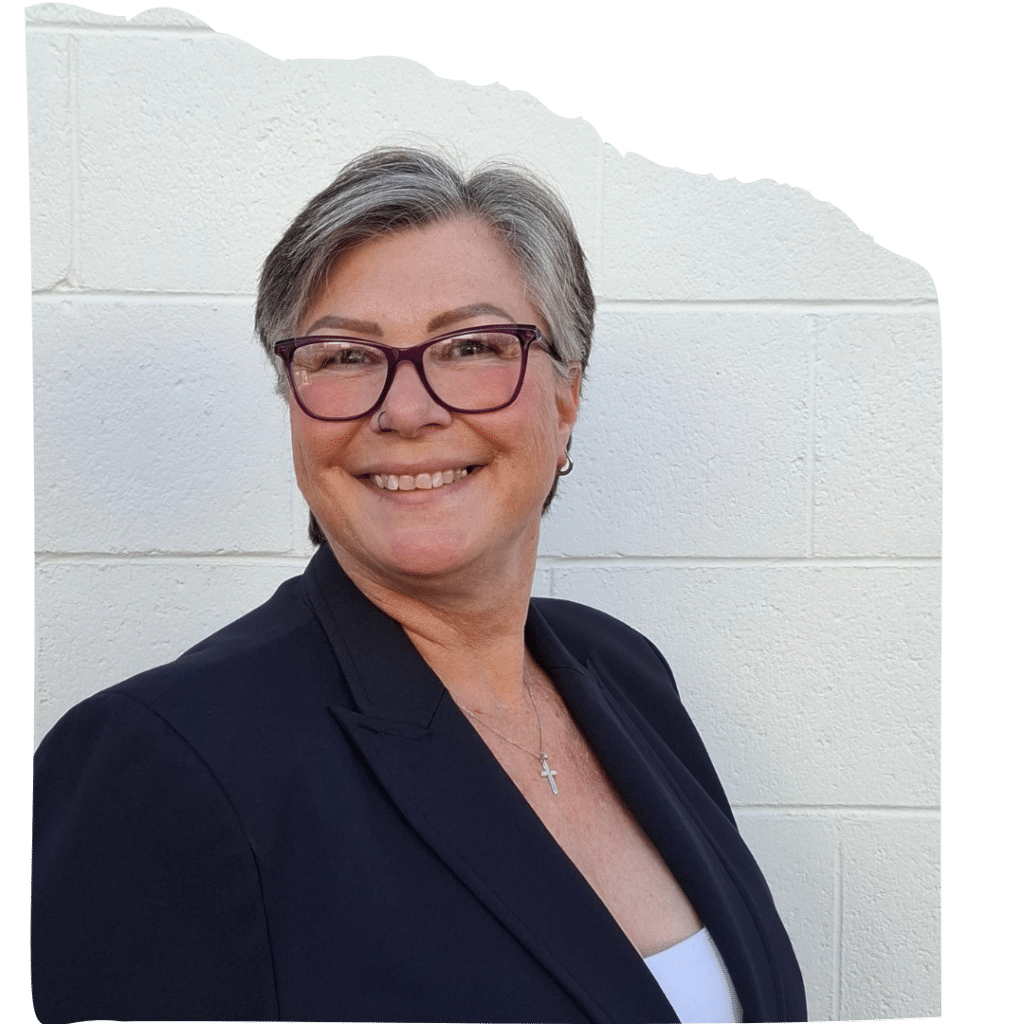 A portrait of Stephanie standing in front of a white brick wall, smiling and wearing a dark blazer, white top, and purple‑framed glasses.